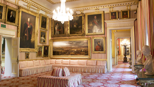 Opulent gallery room of regal figures in historical settings with red and white striped wallpaper, gold-trimmed walls and ceilings with a large chandelier, classical busts and striped sofa
