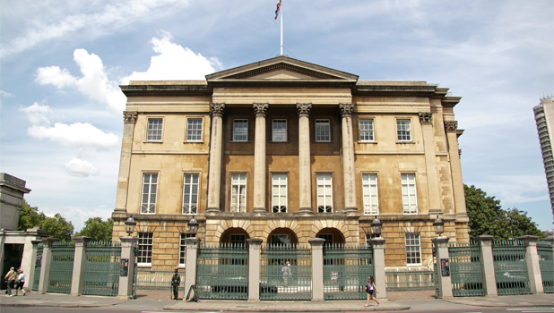 Front view of Aplsey House, a grand classical style building with symmetrical architecture, rectangular windows and a flagpole on top. A green metal fence surrounds the building as people walk by on the street. 