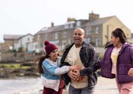 A father and two daughters walking on a beach whilst talking and laughing