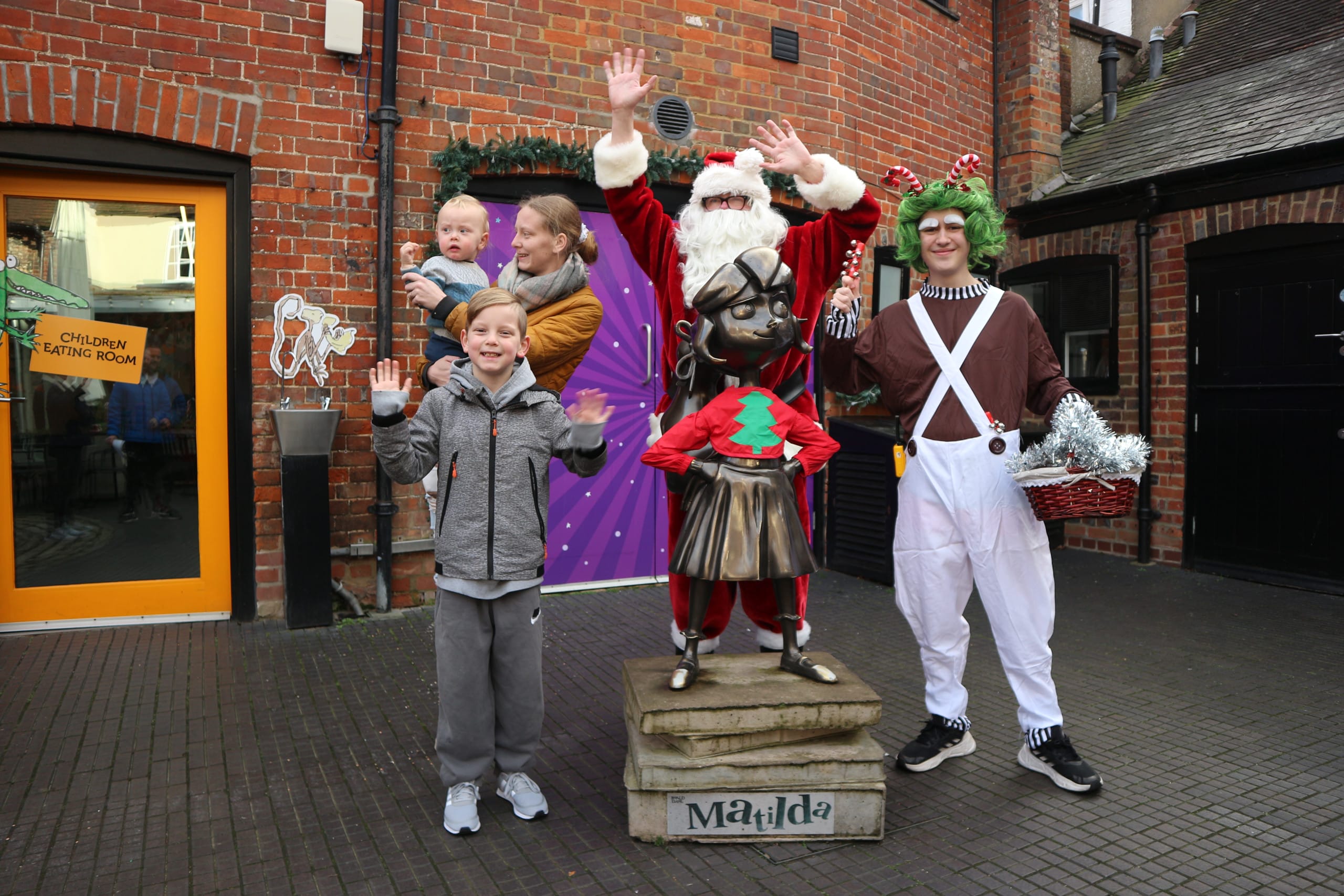 A Matilda statue at the Roald Dahl Museum wearing a red Christmas jumper with a tree. Behind the statue there is a Santa Claue  and a reindeer dressed person plus a mother with an infant child in their arms and a older child standing in front of them
